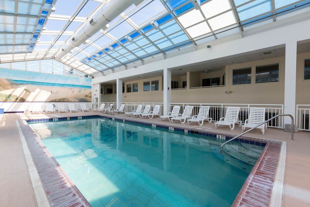 An indoor swimming pool with clear blue water, surrounded by white lounge chairs and a glass ceiling.