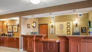 Zona de recepción de un hotel con mostrador, un reloj, premios enmarcados y un cartel que dice 'Comfort Inn & Suites'.