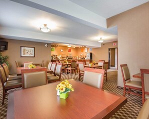 A hotel dining area with a large wooden table in the foreground, surrounded by chairs. There is a buffet counter in the background with various food items and a clock on the wall.