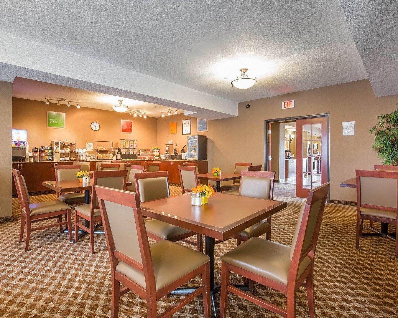 A hotel dining area with multiple tables and chairs, a coffee station, and an exit sign.