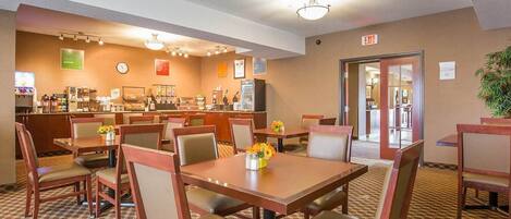A hotel dining area with multiple tables and chairs, a coffee station, and an exit sign.