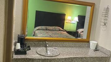A hotel bathroom with a granite countertop, a sink, a mirror, a green wall, a bed with patterned bedding, and a green trash bin.