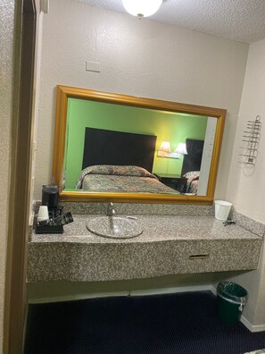 A hotel bathroom with a granite countertop, a sink, a mirror, a green wall, a bed with patterned bedding, and a green trash bin.
