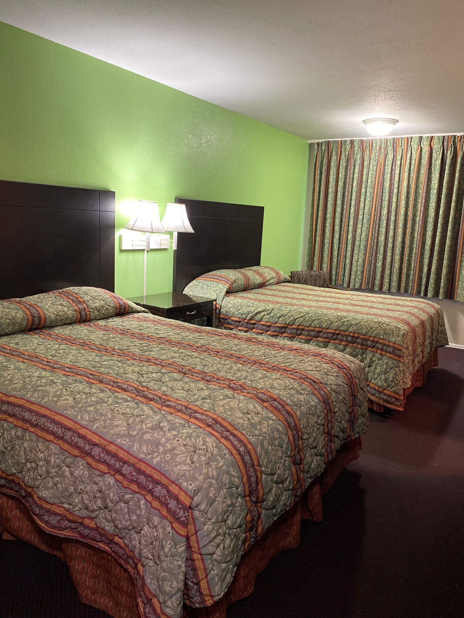 Una habitación de hotel con dos camas, paredes verdes, un cabecero, una mesita de noche con una lámpara y una cortina.