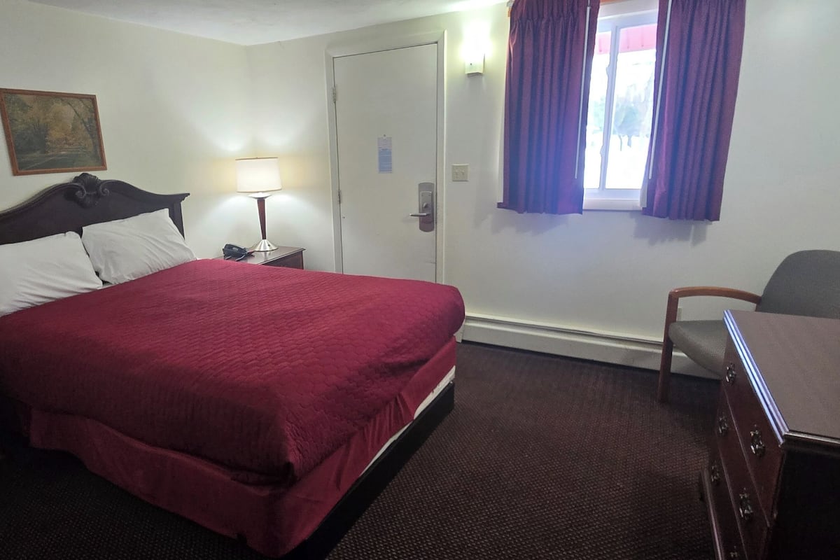 A hotel room with a bed, a desk, a chair, a nightstand, a lamp, and a window with curtains.
