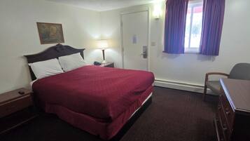 A hotel room with a bed, a desk, a chair, a nightstand, a lamp, and a window with curtains.