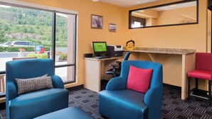 Quarto de hotel com duas poltronas azuis, uma cadeira vermelha, uma secretária com computador e uma janela grande que dá para um estacionamento e áreas verdes.
