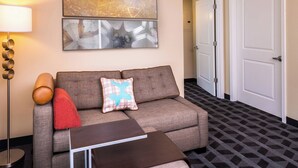 Quarto de hotel moderno com um sofá cinzento, uma mesa pequena, uma luminária e um tapete estampado.