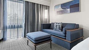 Una habitación de hotel moderna con sofá, reposapiés y un gran ventanal con cortinas.
