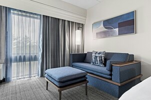 Quarto de hotel moderno com sofá, banquinho e uma janela grande com cortinas.