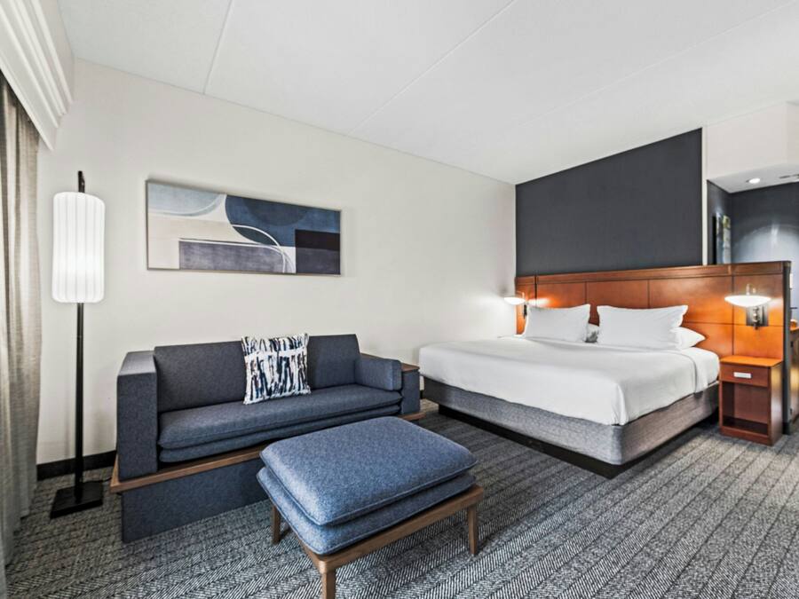 A hotel room with a bed, a sofa, a coffee table, a lamp, a painting, and a nightstand.