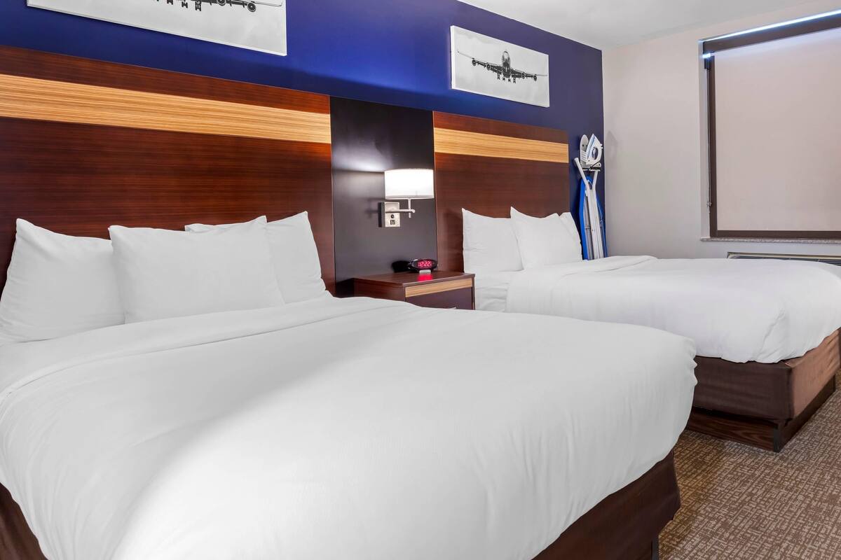 A hotel room with two beds, a nightstand with a lamp, a clock, and a projector screen.