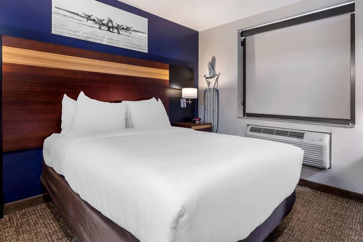 A hotel room with a large bed, a headboard with a wooden panel, a wall-mounted projector screen, a nightstand with a lamp, and a wall-mounted air conditioning unit.