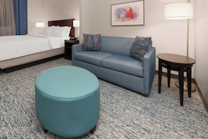 A hotel room with a bed, a sofa, an ottoman, a small table, and a lamp. - Garner Hotel Norman - University Area by IHG (Norman)