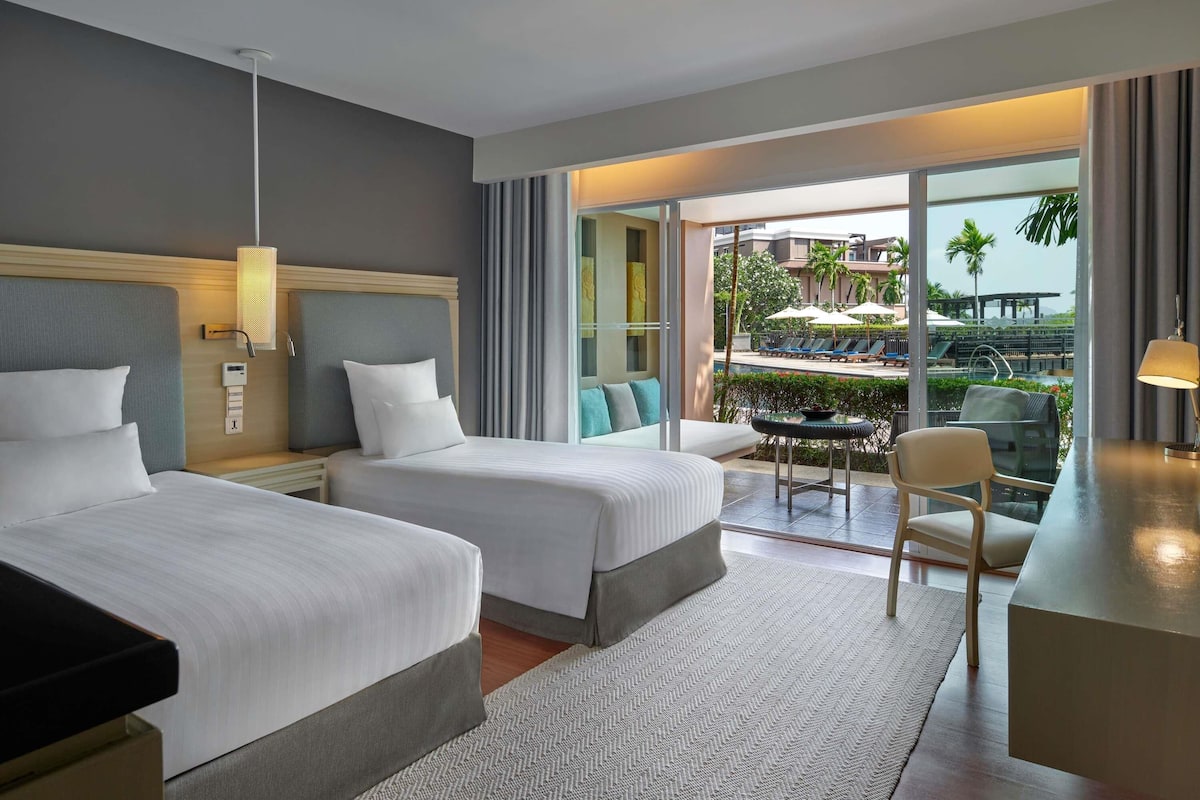 Een hotelkamer met twee bedden, een bureau en uitzicht op een zwembad en palmbomen.