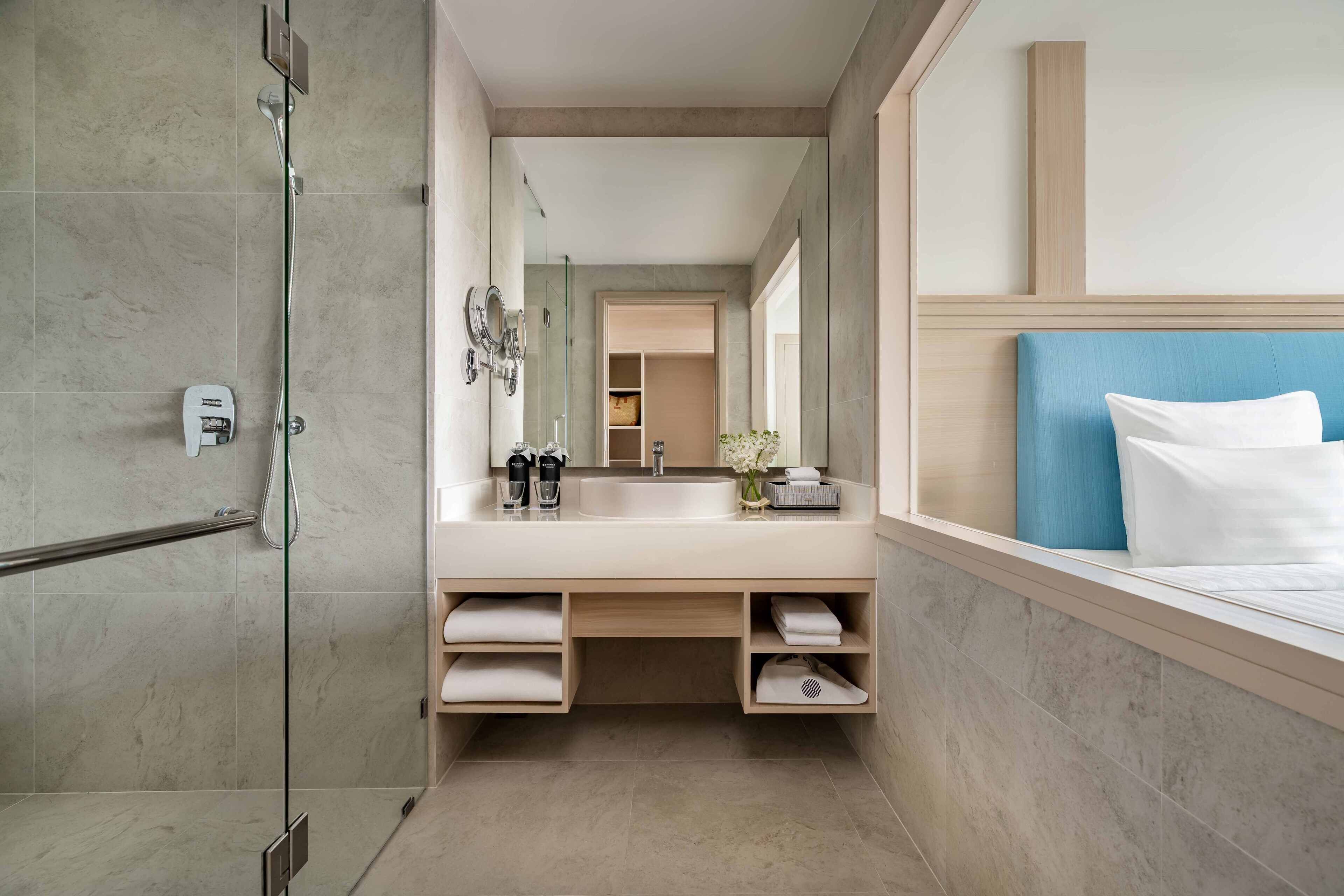 A modern bathroom with a glass shower enclosure, a large mirror, a sink with a towel rack, and a bed with a blue headboard.