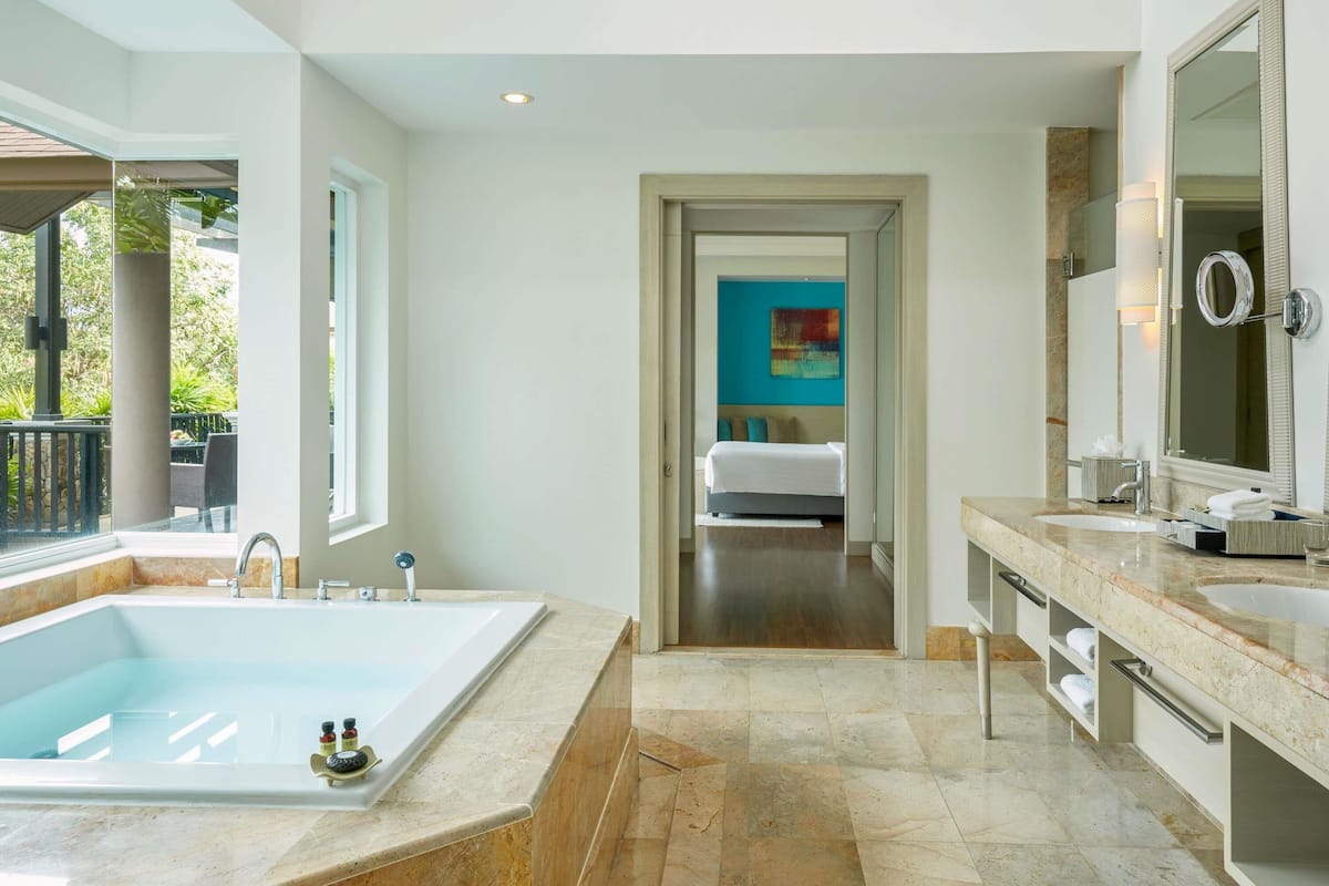 Ein modernes Badezimmer mit einer großen Badewanne, einer begehbaren Dusche und einem angrenzenden Schlafzimmer, das durch eine offene Tür sichtbar ist.