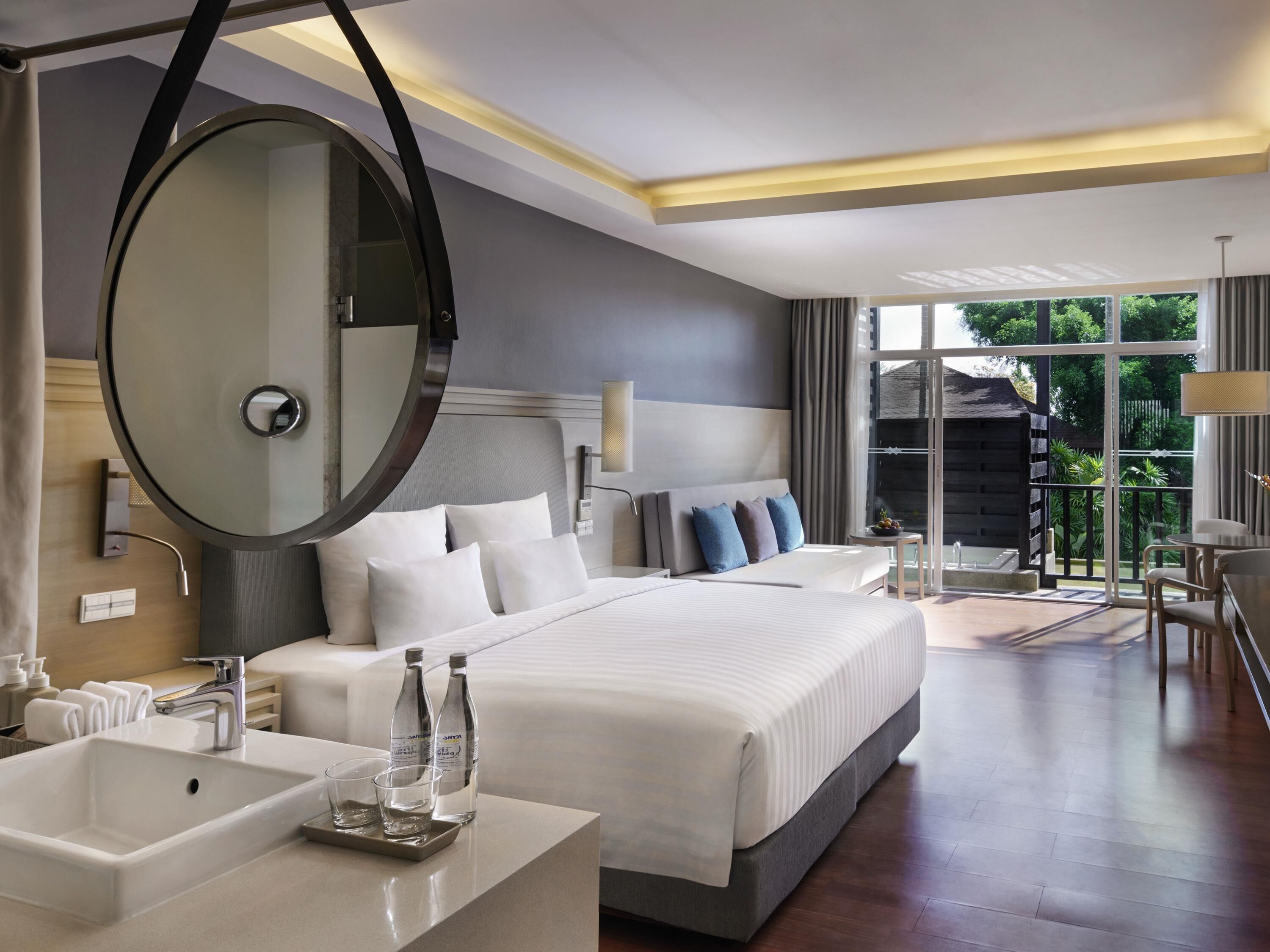 Une chambre d’hôtel moderne avec un grand lit, un miroir rond, une table de chevet dotée d’un lavabo et de verres, et une vue sur l’extérieur.