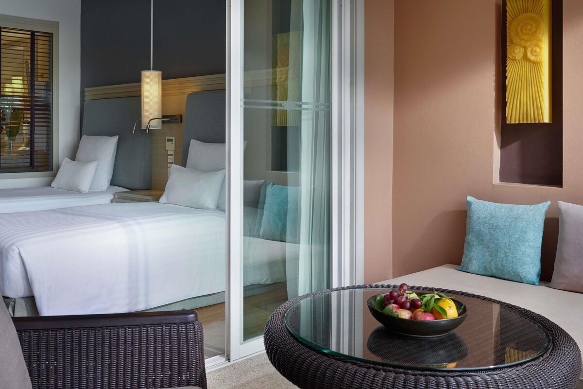 Una habitación de hotel moderna con una cama, un vidrio que da a un balcón, una mesa redonda de vidrio con fruta, y un sofá con cojines.