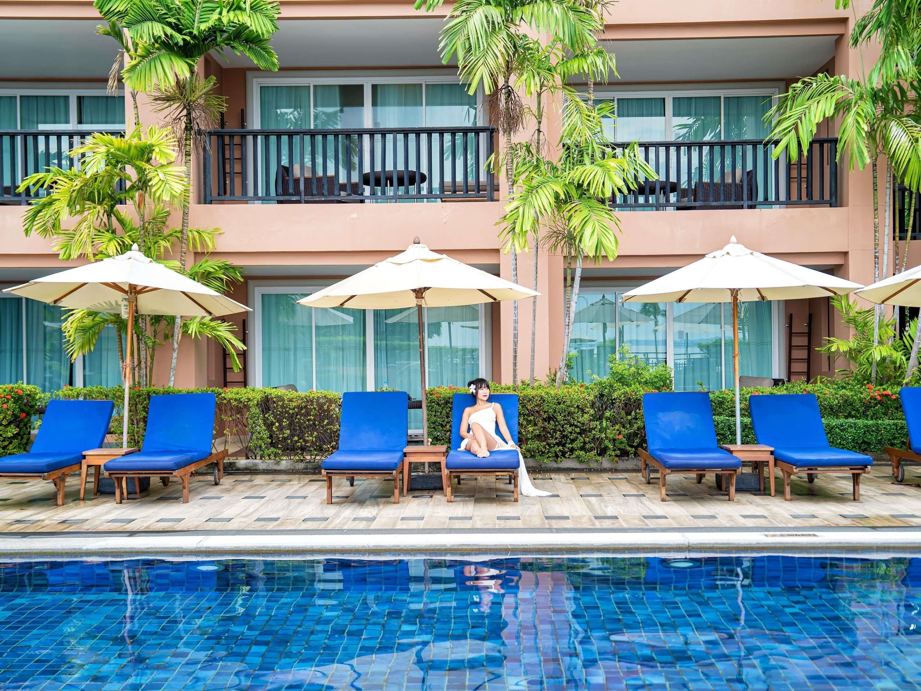 Un espace aménagé au bord de la piscine, avec des chaises longues et des parasols, une piscine aux carreaux bleus et un bâtiment doté de balcons et de fenêtres.