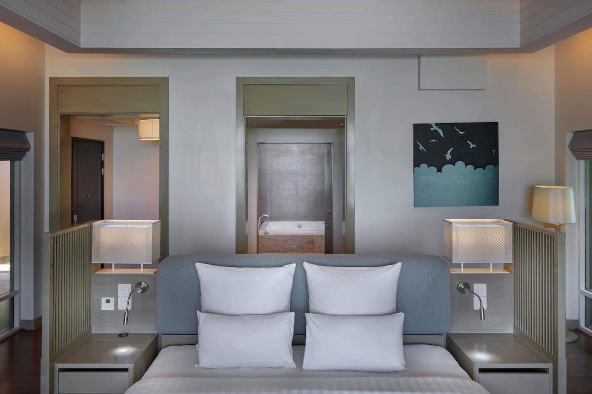 A hotel room with a large bed, two bedside tables with lamps, a mirror, a painting, and a view of the bathroom.