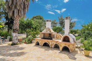 An outdoor stone barbecue area with a chimney, surrounded by greenery and a clear sky. - Villas Therianos (Zakynthos)