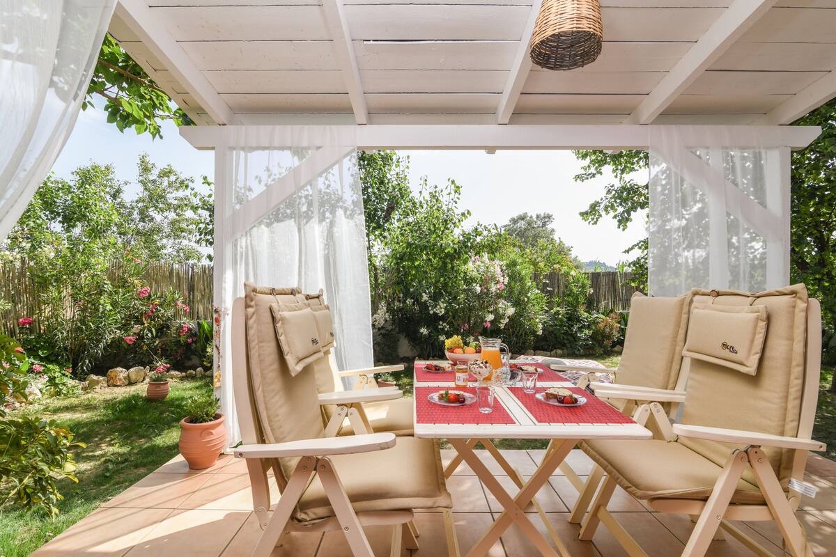 An outdoor patio with a white pergola, two beige cushioned chairs, a table set with plates, glasses, and a pitcher of orange juice.
