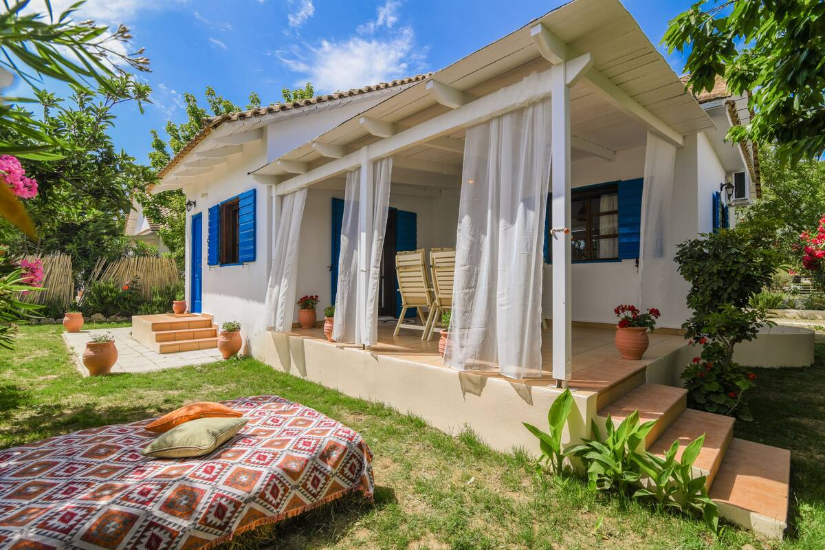 A white house with blue shutters, a covered porch, and a garden with potted plants and a bed.