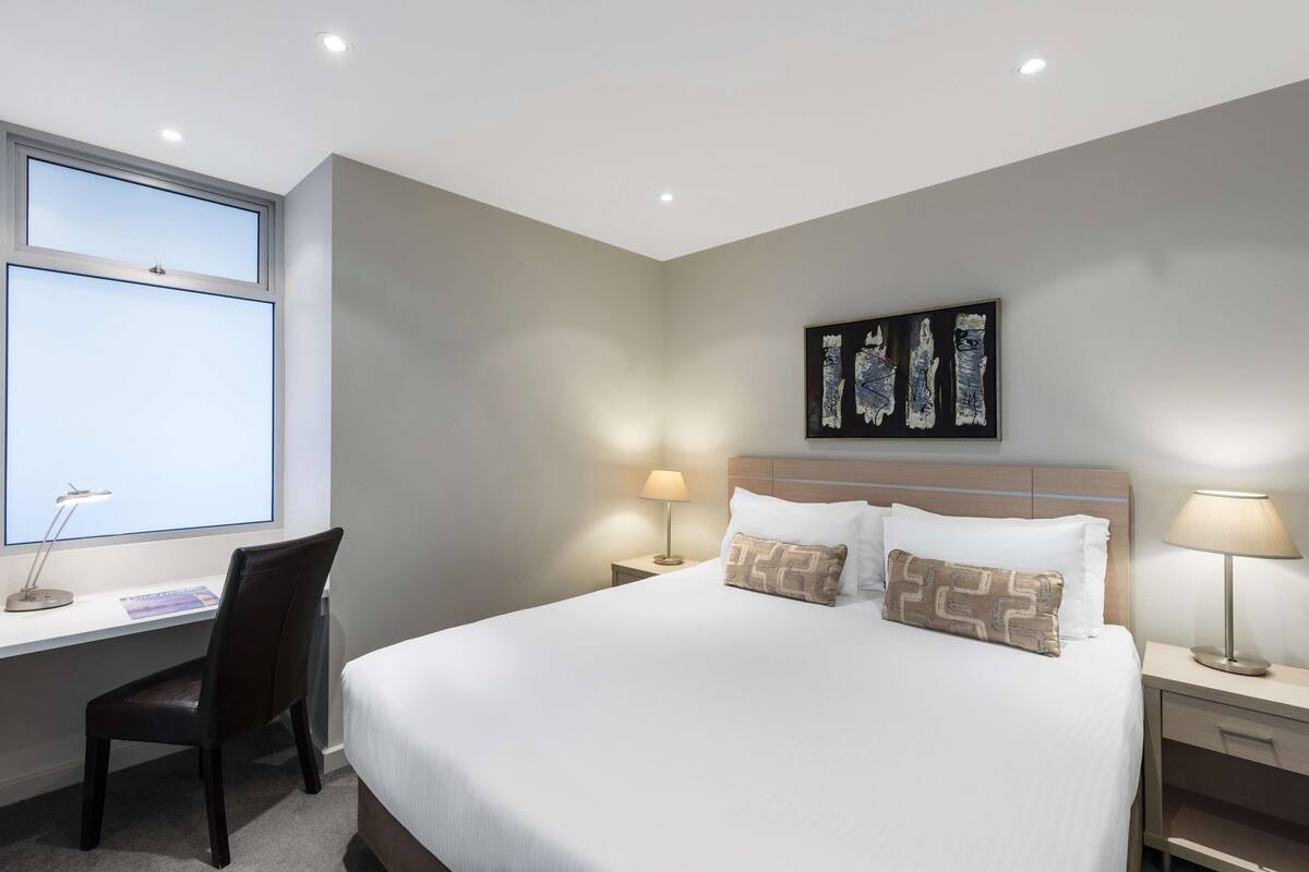Habitación de hotel con cama, escritorio, silla, dos lámparas de noche y un cuadro en la pared.