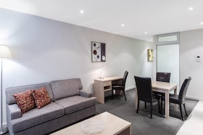 A modern living room with a grey sofa, a small table, a desk with a chair, and a dining table with chairs.