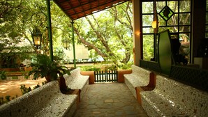 A covered outdoor seating area with mosaic-tiled benches and a stone floor, surrounded by greenery and a view of trees. - Indeco Mahabalipuram (Tirukalukundram)