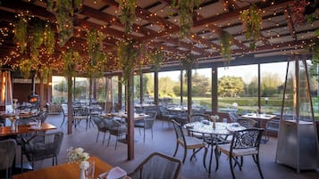 Un espacio de comedor al aire libre con mesas y sillas, luces colgantes y vistas al exterior.