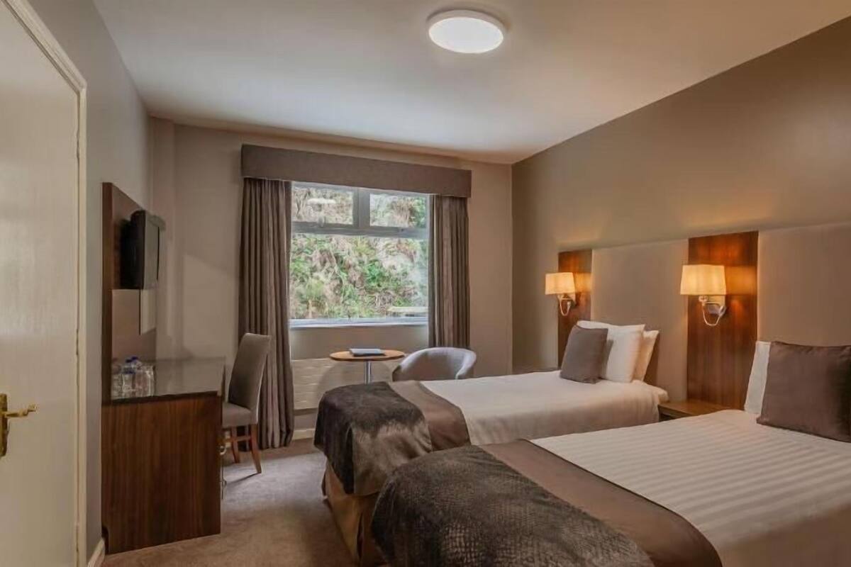 Une chambre d’hôtel avec deux lits, un bureau, une chaise, une fenêtre avec des rideaux et des lampes fixées au mur.