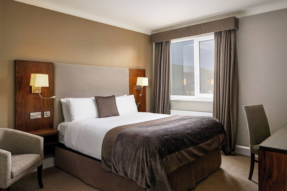 Une chambre d’hôtel avec un grand lit, deux lampes de chevet, une chaise, un bureau et une fenêtre avec des rideaux.