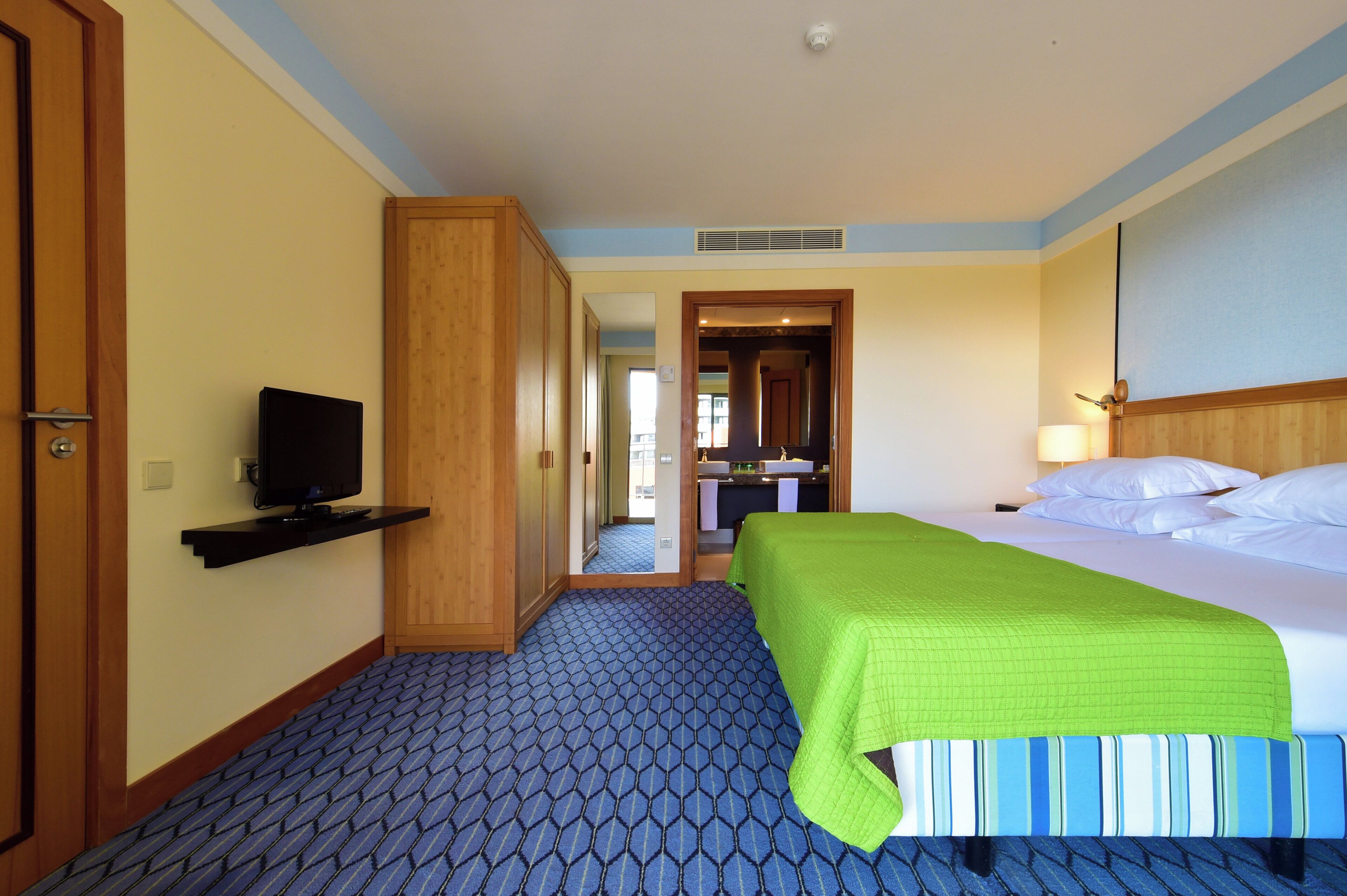 Hotelkamer met een bed, televisie, badkamer met wastafel en spiegel, en een gang naar een andere kamer.