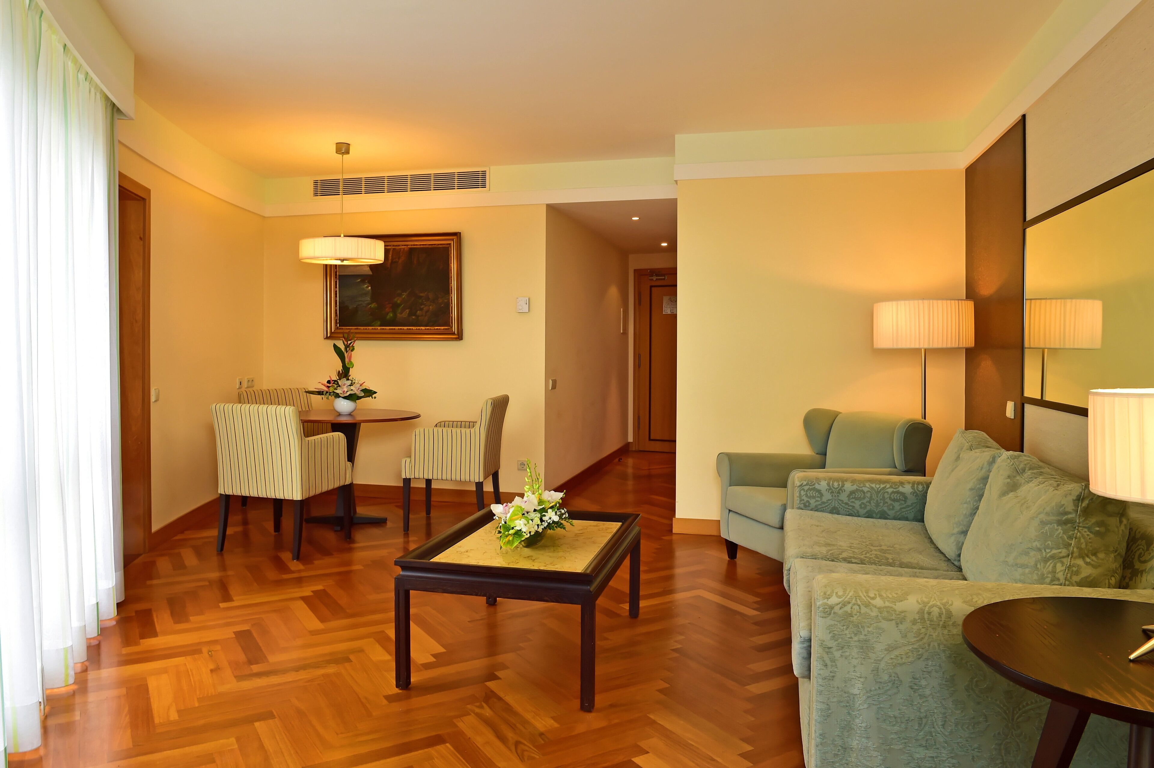 A hotel room with a wooden parquet floor, a dining table with chairs, a sofa, and a lamp.