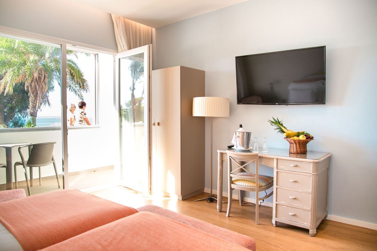 A hotel room with a bed, a desk with a lamp, a TV, and a view of palm trees.