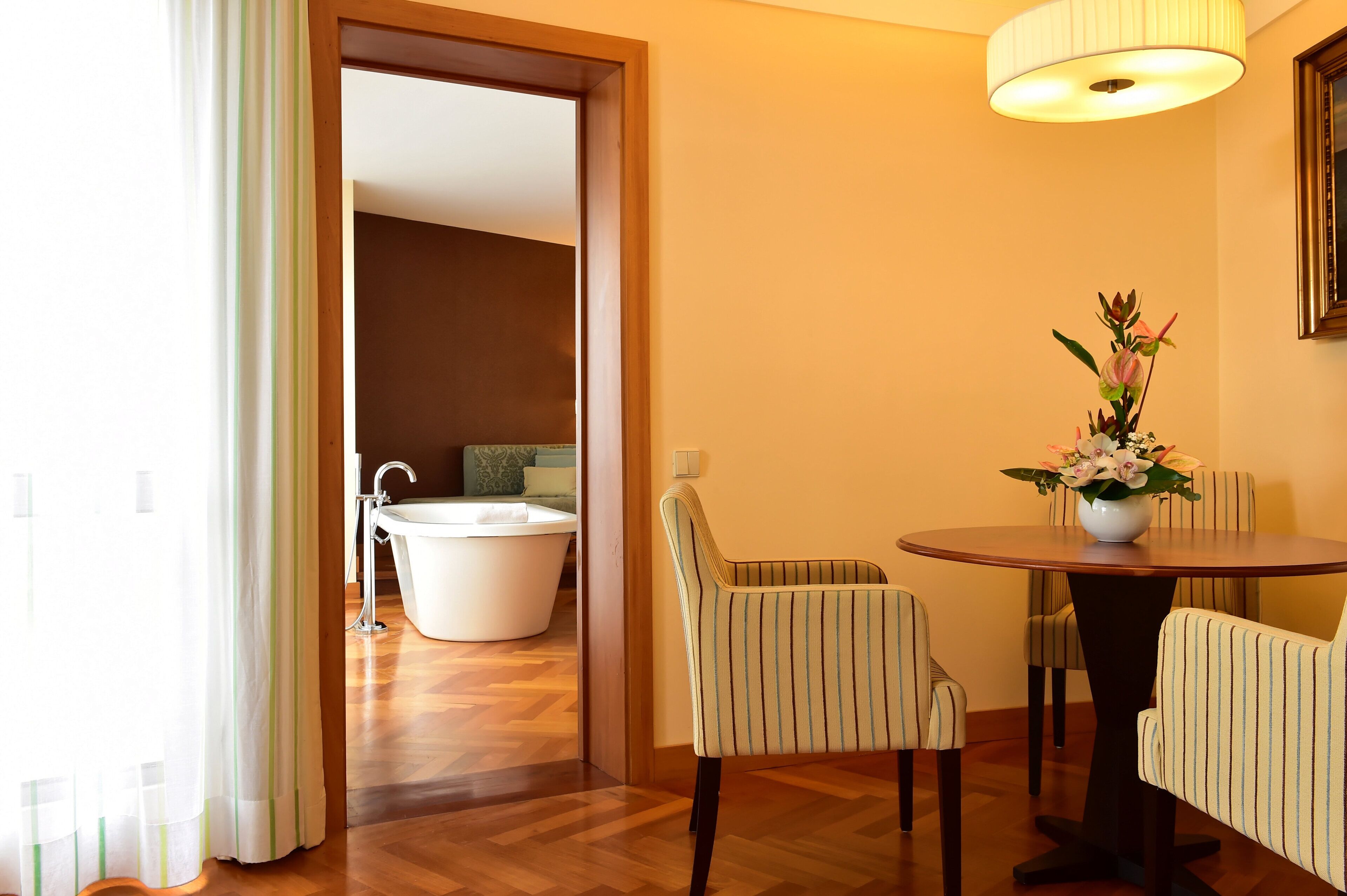 Una habitación de hotel con piso de madera, una mesa redonda de comedor con sillas a rayas, una bañera blanca y una planta en maceta.