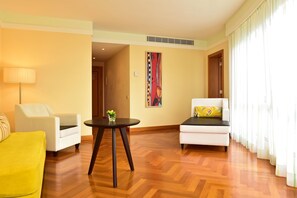 Habitación de hotel con piso de madera, dos sillones blancos, un sillón amarillo, una mesita redonda con una planta y un cuadro en la pared.