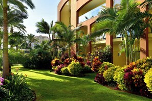 Un jardín bien cuidado con palmeras y plantas coloridas frente a un edificio amarillo con balcones.