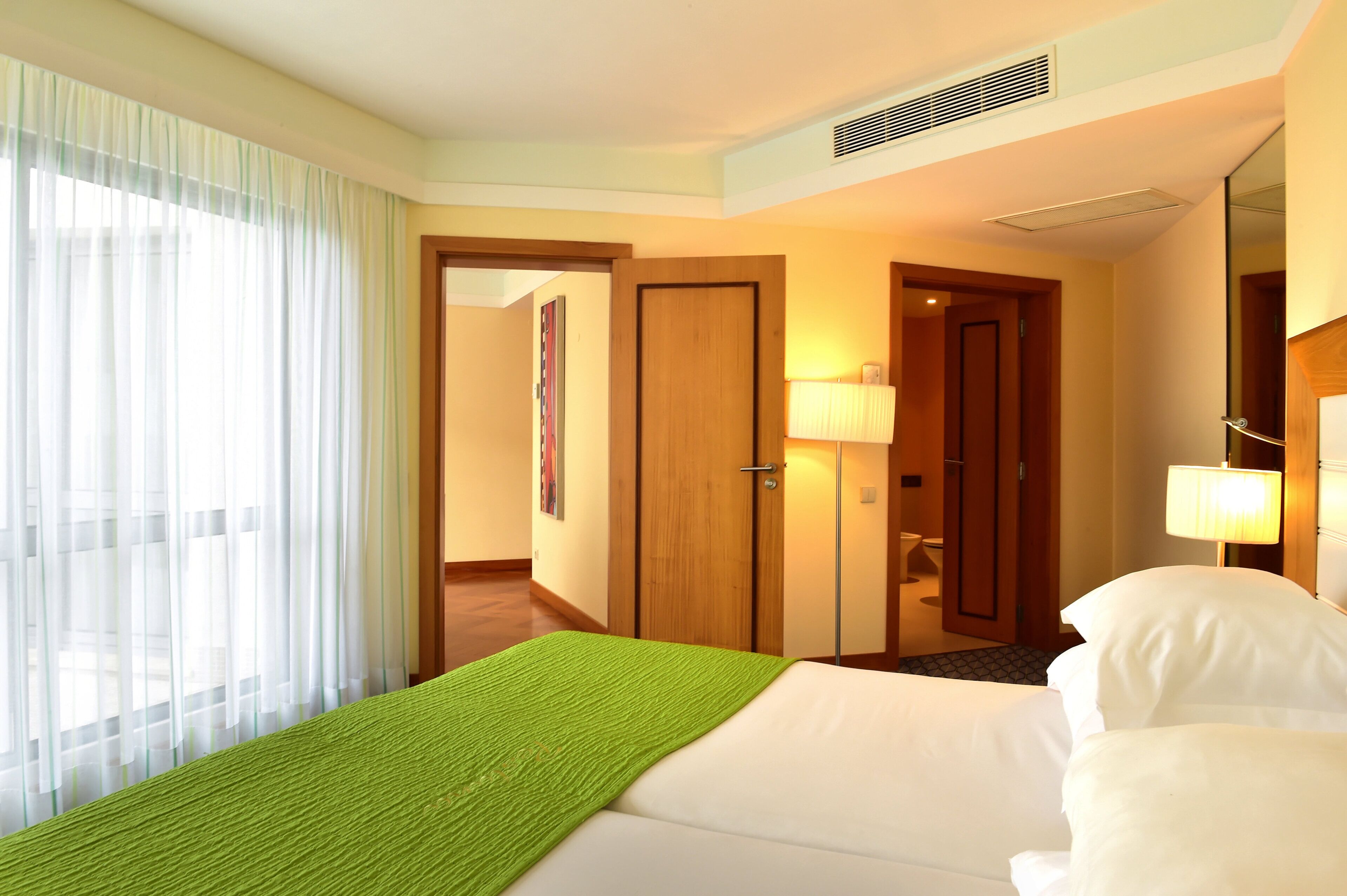 Une chambre d’hôtel avec un lit, une fenêtre avec des rideaux légers, une porte et un couloir menant à une salle de bain.