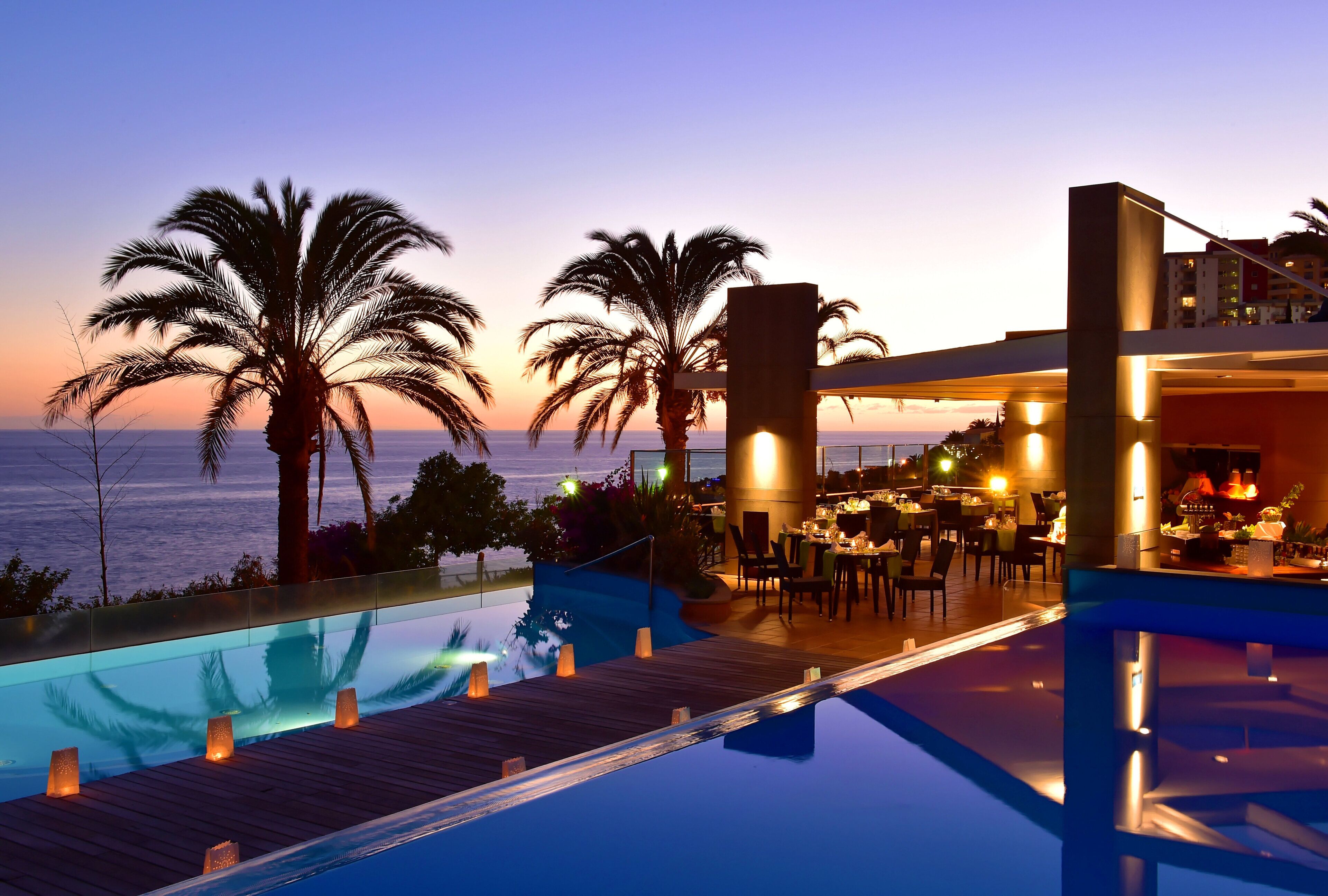 Zona junto a la piscina con vista al océano, palmeras y un área de comedor con mesas y sillas.