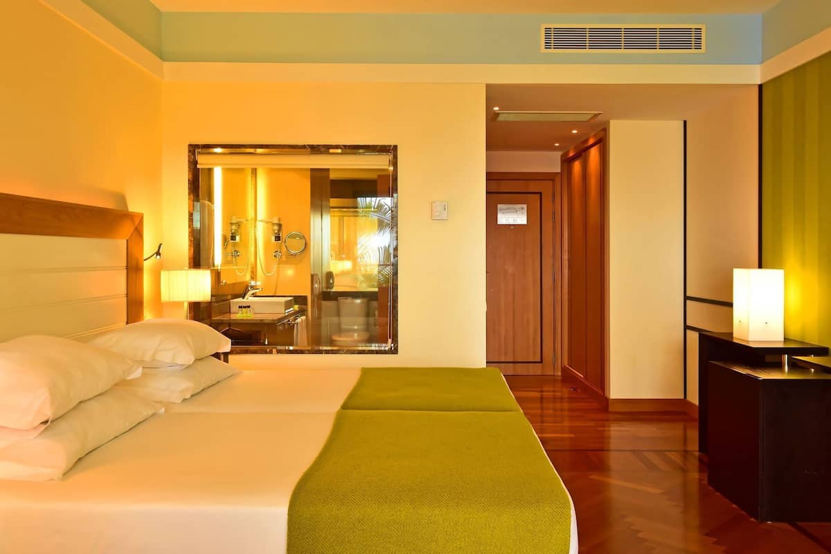 A hotel room with a bed, a green throw, a nightstand, a lamp, and a view into the bathroom.