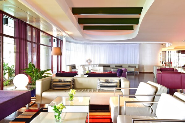 Um hall de hotel moderno com teto curvo, área de estar com sofás e poltronas, mesa de centro com um vaso e uma grande janela com cortinas.