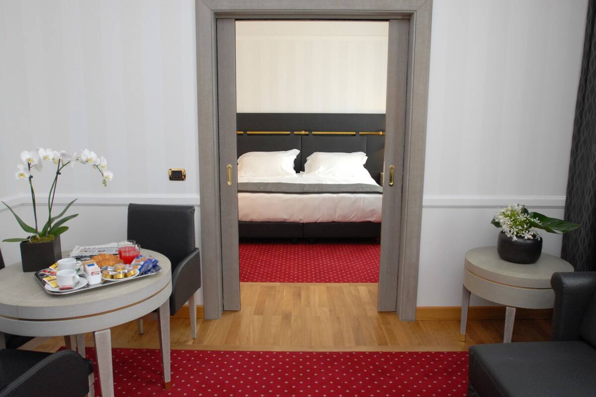 Una habitación de hotel con cama, zona de comedor y una bandeja de desayuno, junto con dos plantas en macetas.