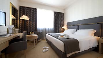 A hotel room with a bed, desk, chair, and a small table with a lamp.