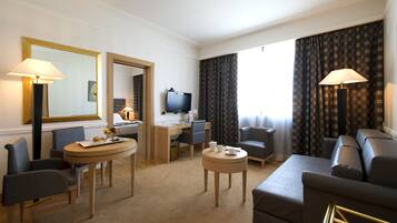 Habitació d'hotel amb zona de menjador, televisió, escriptori, sofà i finestra gran amb cortines.