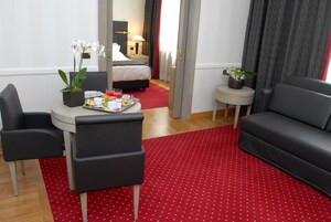 Una habitación de hotel con una cama, una mesita con una bandeja de desayuno, dos sillones, una alfombra roja y una planta en maceta.