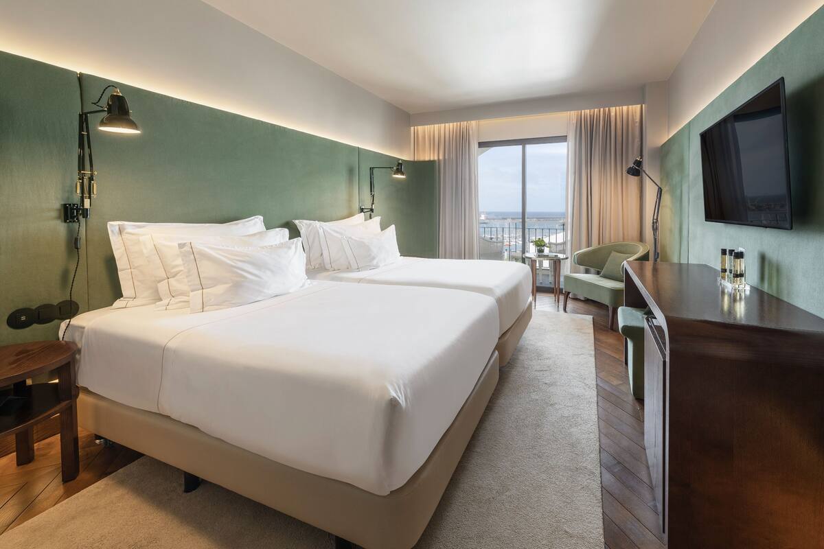 Een hotelkamer met twee bedden, een bureau, een stoel, een televisie en uitzicht op de stad.