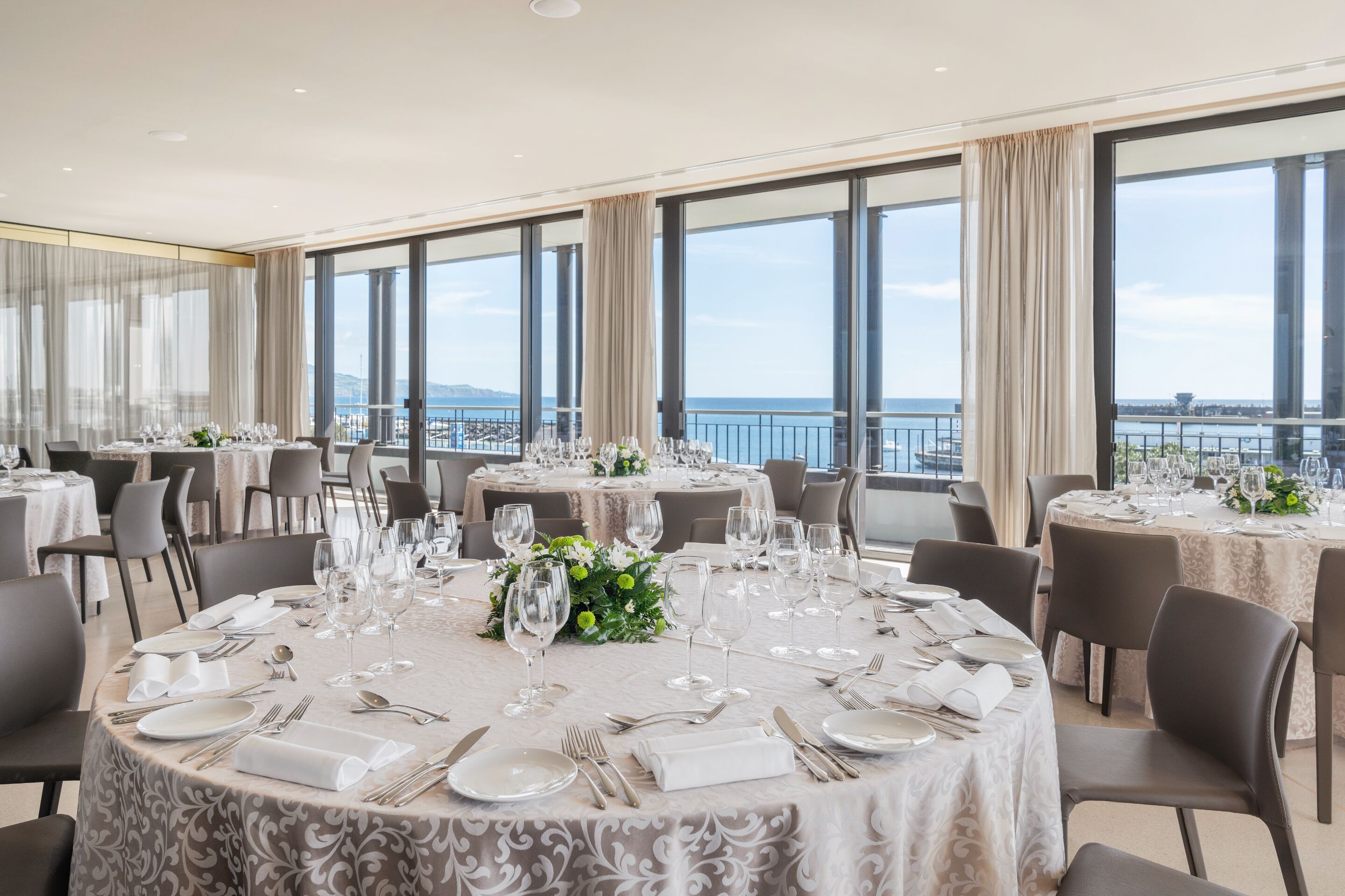 A dining area with round tables set for a formal meal, large windows offering a view of the sea, and a balcony with a railing.